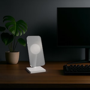 Può includere: Un supporto di ricarica wireless bianco caratterizzato da un pannello verticale rettangolare e traslucido con un'area di ricarica magnetica circolare al centro, montato su una base quadrata, progettato per sostenere uno smartphone in verticale.