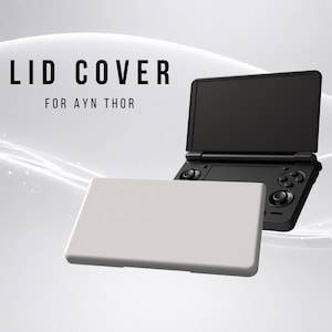 May include: A solid light gray rectangular lid cover for the Ayn Thor handheld gaming console. The cover is positioned in front of a black handheld gaming device with a screen and control buttons. The text LID COVER FOR AYN THOR is visible.
