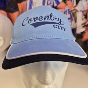May include: A light blue baseball cap with a white brim and a navy blue underside. The cap has a navy blue logo that says "Coventry City" in a cursive font.