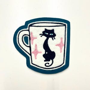 May include: Embroidered patch in the shape of a coffee mug. The mug is white with a black cat silhouette and pink starbursts. The mug is outlined in black and has a teal border. The patch is likely for a jacket, bag, or other fabric item.