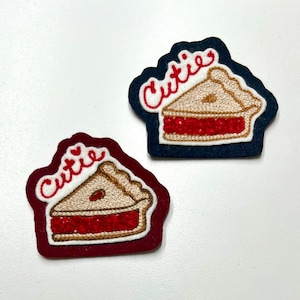 May include: Two embroidered iron-on patches with a pie slice design. Both patches have the word "Cutie" embroidered on them. One patch is on a dark blue felt background and the other is on a burgundy felt background.