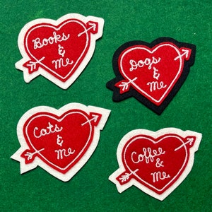May include: Four felt heart-shaped patches with white embroidery. Each patch features an arrow and the text "Books & Me", "Dogs & Me", "Cats & Me", and "Coffee & Me".