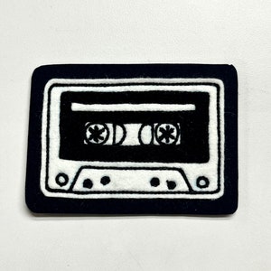May include: Black and white embroidered cassette tape patch. The patch is rectangular and has a black border. The cassette tape is white with black details.