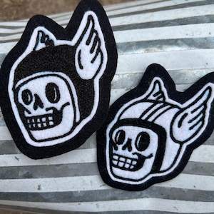 May include: Two black and white embroidered patches. Each patch features a skull wearing a winged helmet. The skull has a wide grin and is outlined in white. The patches are on a corrugated metal surface.