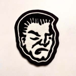 Peut inclure: Patch en feutre noir et blanc représentant un visage d'homme avec une expression sérieuse. Le visage est délimité par un contour noir et les détails sont brodés en blanc.
