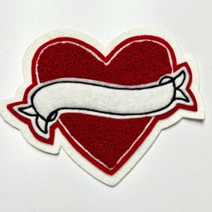 May include: Red felt heart with a white felt banner, outlined in black, with a simple design. The banner is blank.