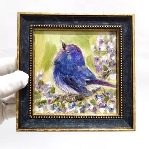 May include: A framed oil painting of a blue bird perched on a branch with purple and white flowers. The bird is painted with shades of blue and purple. The frame is dark blue with a gold inner border.