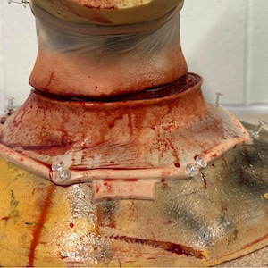 Pode incluir: Um pescoço protético com um corte simulado, apresentando um tom de pele realista e uma substância semelhante a sangue. Pinos transparentes prendem as bordas da prótese a uma superfície amarela e marrom, criando um efeito horripilante.