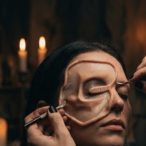 May include: A person's face with a prosthetic eye and brow piece being applied. The prosthetic is flesh-toned with the word "KORY" printed on it. The person's eyes are closed. The background is dark with two lit candles.