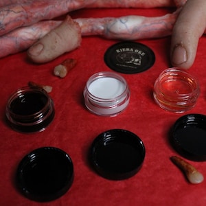 Może przedstawiać: Asortyment słoiczków z czarnymi wieczkami, zawierających różne substancje, wraz z kilkoma zębami i palcem. Słoiczki są przezroczyste, ukazując zawartość, i ułożone na czerwonej powierzchni. Obecna jest również czarna okrągła etykieta z białym tekstem.