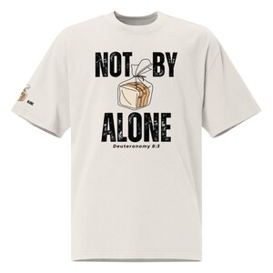 Op de afbeelding: Crèmekleurig T-shirt met de tekst "NOT BY ALONE" boven een illustratie van een brood in een plastic zak. De tekst "Deuteronomium 8:3" staat onder "ALONE". Een kleine broodillustratie staat op de linkermouw.