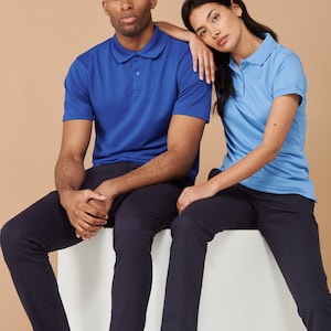 K&ouml;nnte beinhalten: Zwei Personen sitzen auf einer wei&szlig;en Fl&auml;che. Die Person links tr&auml;gt ein blaues Poloshirt und eine dunkelblaue Hose. Die Person rechts tr&auml;gt ein hellblaues Poloshirt und eine dunkelblaue Hose. Beide tragen wei&szlig;e Turnschuhe.