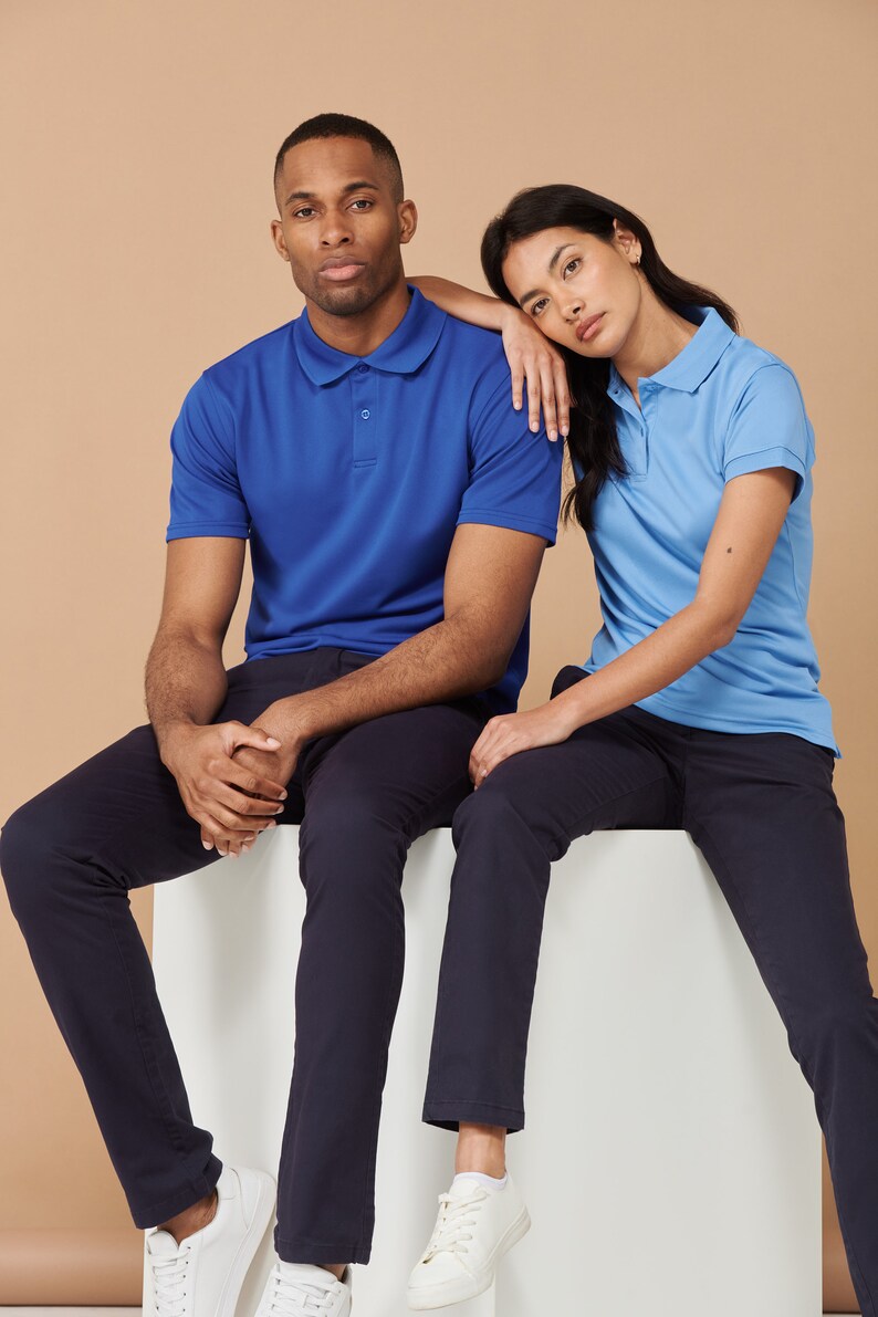 K&ouml;nnte beinhalten: Zwei Personen sitzen auf einer wei&szlig;en Fl&auml;che. Die Person links tr&auml;gt ein blaues Poloshirt und eine dunkelblaue Hose. Die Person rechts tr&auml;gt ein hellblaues Poloshirt und eine dunkelblaue Hose. Beide tragen wei&szlig;e Turnschuhe.
