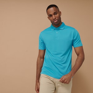 K&ouml;nnte beinhalten: Ein Mann tr&auml;gt ein hellblaues Poloshirt mit einer Zwei-Knopf-Leiste. Er steht vor einem beigefarbenen Hintergrund.