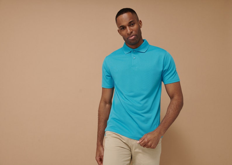K&ouml;nnte beinhalten: Ein Mann tr&auml;gt ein hellblaues Poloshirt mit einer Zwei-Knopf-Leiste. Er steht vor einem beigefarbenen Hintergrund.