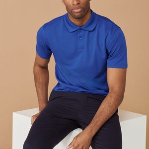 K&ouml;nnte beinhalten: Ein Mann in einem blauen Poloshirt und einer dunkelblauen Hose sitzt auf einem wei&szlig;en W&uuml;rfel. Er schaut in die Kamera.