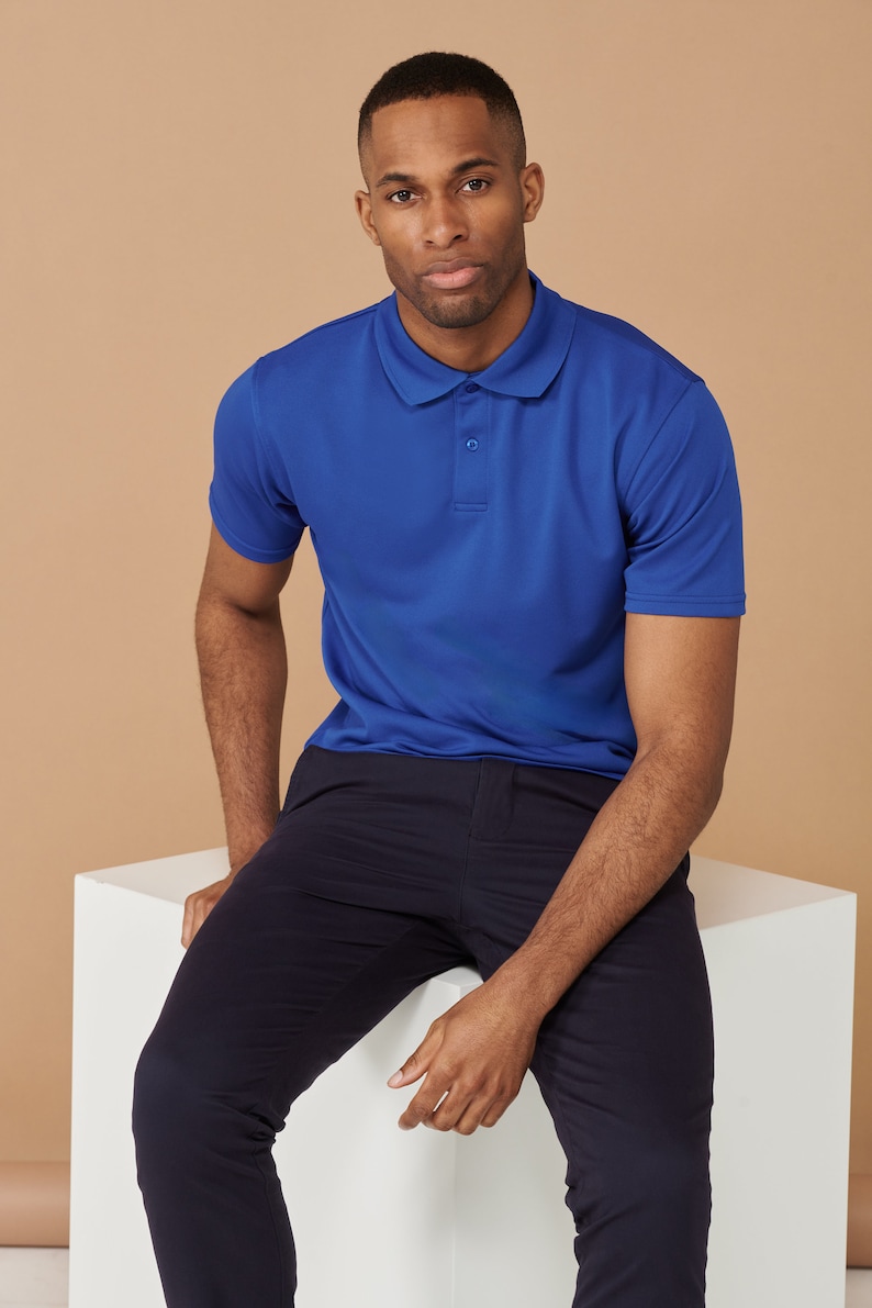 K&ouml;nnte beinhalten: Ein Mann in einem blauen Poloshirt und einer dunkelblauen Hose sitzt auf einem wei&szlig;en W&uuml;rfel. Er schaut in die Kamera.
