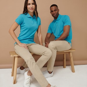 K&ouml;nnte beinhalten: Zwei Personen sitzen auf einer Holzbank. Sie tragen beide hellblaue Poloshirts und Khaki-Hosen. Die Frau sitzt auf der linken Seite der Bank und der Mann sitzt auf der rechten Seite der Bank. Beide schauen in die Kamera.