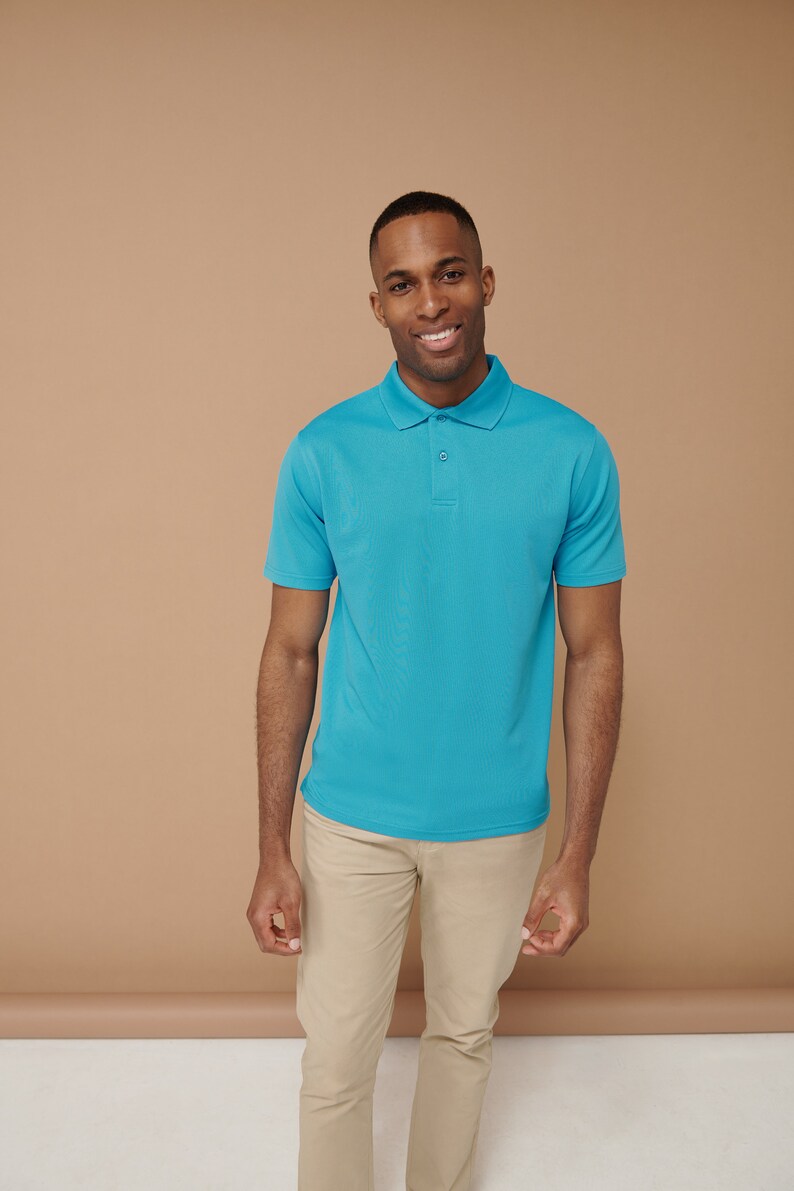 K&ouml;nnte beinhalten: Ein Mann tr&auml;gt ein hellblaues Poloshirt und eine Khakihose. Er l&auml;chelt und schaut in die Kamera.