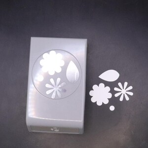May include: A grey paper punch with four different shapes: a large flower, a small flower, a leaf, and a circle. The punch is on a grey surface and there are four punched out shapes next to it.