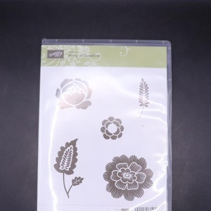 Può includere: Una confezione di plastica trasparente contenente un set di cinque timbri in gomma trasparente con motivi floreali e di foglie. La confezione è etichettata "Hostess Array of Sunshine" e presenta il logo Stampin' Up!