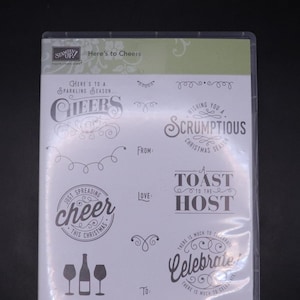 Pode incluir: Uma embalagem de plástico transparente contendo um conjunto de 13 carimbos de borracha com tema natalino. Os carimbos apresentam várias saudações e designs de férias, incluindo "Cheers", "Scrumtious Christmas Season", "A Toast to the Host", e "Celebrate".