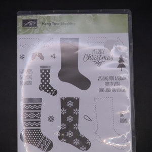 Puede incluir: Un paquete de plástico transparente que contiene un conjunto de 19 sellos de goma con temática navideña. Los sellos presentan una variedad de diseños, incluyendo calcetines, copos de nieve, acebo y las palabras "Feliz Navidad" y "¡La felicidad llega a la ciudad!" El paquete también incluye un sello de sentimiento que dice "Te deseo una temporada llena de amor y felicidad." y un sello "Para: De: "