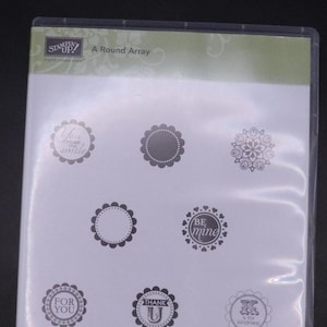 Può includere: Una confezione di plastica trasparente contenente un set di otto timbri in gomma. I timbri presentano varie frasi e disegni, tra cui "You make me smile", "Be mine", "For you", "Thank U" e "K is for kindness". La confezione include anche il testo "A Round Array" e "Set of 8".