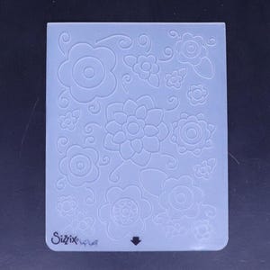 May include: A white silicone embossing mat with a floral design. The mat has a Sizzix logo in the bottom left corner.