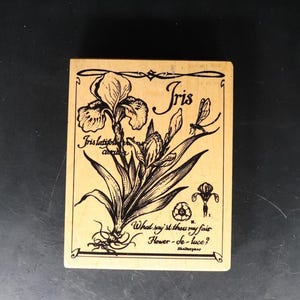 May include: A wooden stamp featuring a black and white illustration of an iris flower and a dragonfly. The text "Iris" is printed above the flower and "Iris latifolia" is printed below the flower. The text "What say'st thou my fair Flower-de-luce?" is printed below the flower.