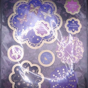 Op de afbeelding: Een set paarse en gouden decoratieve papieren kaarten met vlinderontwerpen. De kaarten bevatten verschillende berichten, waaronder "Come Very Special", "Just For You", "Thinking of You" en "Happy Birthday".