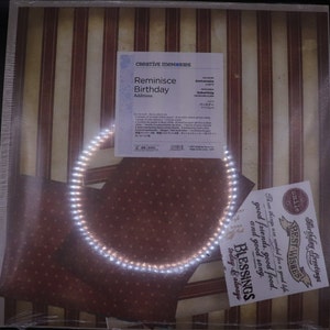 Könnte beinhalten: Eine Packung Creative Memories Reminisce Birthday Additions Scrapbooking-Zubehör. Die Packung enthält gemustertes Papier, Aufkleber und Verzierungen. Die Packung befindet sich auf einem rot-weiß gestreiften Hintergrund mit einem weißen Lichtkreis.