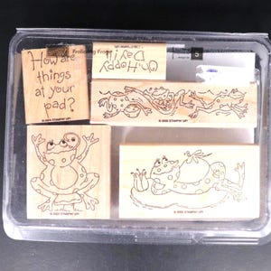 Peut inclure: Un ensemble de tampons en bois avec des illustrations de grenouilles et des phrases. Les tampons incluent une grenouille avec les bras en l'air, une grenouille sur un nénuphar et un tampon avec le texte "How are things at your pad?"