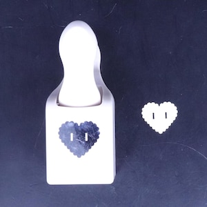 Puede incluir: Perforadora de papel de plástico blanco en forma de corazón con un diseño de corazón negro. La perforadora se muestra junto a una muestra de un corazón perforado.
