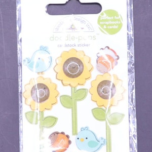 Op de afbeelding: Een pakket "doodle-pops" kartonnen stickers. De stickers tonen cartoon zonnebloemen, vogels en andere kleine wezens in geel, bruin, oranje en blauw. De verpakking heeft een groene koptekst en de tekst "perfect for scrapbooks & cards!"