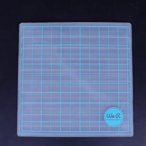 Puede incluir: Una estera de corte de plástico transparente con un patrón de cuadrícula y un logotipo azul We R Memory Keepers en la esquina inferior derecha.
