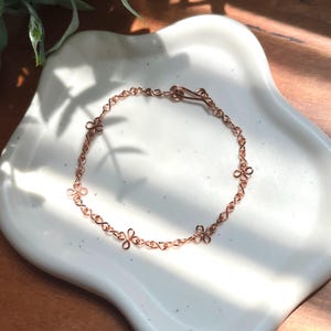 May include: A rose gold-toned wire bracelet featuring a repeating pattern of small, four-petaled flower motifs linked together by twisted wire segments. The bracelet is secured with a simple hook-and-eye clasp mechanism made from the same wire.