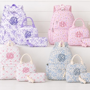 Puede incluir: Una colección de mochilas, bolsas de almuerzo y estuches acolchados con varios estampados florales en morado, rosa, blanco y azul. Cada artículo presenta un bordado con monograma en un color de hilo a juego y un cierre de cremallera.