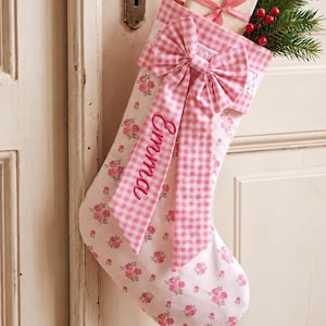 Op de afbeelding: Een roze en witte kerstkous met een bloemenpatroon op het lichaam en een roze geruite boord. Een grote bijpassende geruite strik is aan de voorkant bevestigd, geborduurd met de naam Emma. Een klein cadeautje en dennentakjes zitten erin.