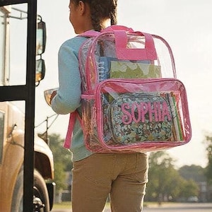 Pu&ograve; includere: Uno zaino trasparente con finiture rosa e il nome "SOPHIA" in lettere rosa sulla tasca anteriore. Lo zaino &egrave; pieno di materiale scolastico. La persona &egrave; vicino a un autobus scolastico.