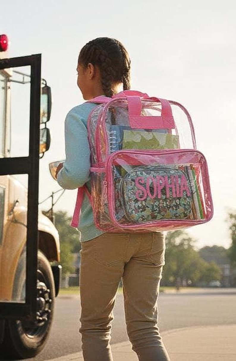 Pu&ograve; includere: Uno zaino trasparente con finiture rosa e il nome "SOPHIA" in lettere rosa sulla tasca anteriore. Lo zaino &egrave; pieno di materiale scolastico. La persona &egrave; vicino a un autobus scolastico.