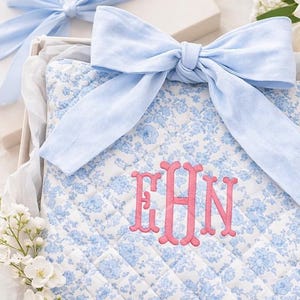 May include: A quilted fabric item featuring a blue and white floral pattern, adorned with a large light blue fabric bow. The center displays the embroidered pink monogram letters EHN. The item is presented in a box with white tissue paper.