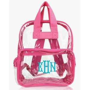 Puede incluir: Una mochila transparente con ribetes, correas y asa superior rosas. El bolsillo delantero presenta las iniciales bordadas "EHN" en color turquesa. La mochila tiene un bolsillo frontal con cremallera y correas ajustables.