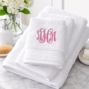 May include: A stack of white cotton towels featuring a pink embroidered monogram on the top towel. The towels have a textured border detail near the folded edge. The embroidery consists of three intertwined cursive letters in a bright pink thread.