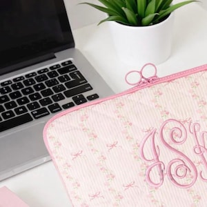 May include: Pink laptop sleeve with a floral and bow pattern. The sleeve has a pink zipper and a monogram in the center. A laptop and a potted plant are in the background.