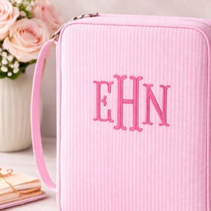 May include: A light pink rectangular zippered case featuring a subtle vertical striped pattern and a matching pink handle. The front of the case is embroidered with the monogram letters EHN in a bold, dark pink thread centered on the surface.