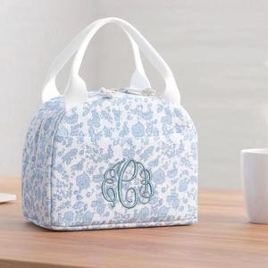 Könnte beinhalten: Eine hellblau-weiß gemusterte Lunchtasche mit weißem Griff und tealfarbenem Monogramm. Die Tasche hat einen Reißverschluss und steht auf einer Holzoberfläche. Die Tasche ist für den Transport von Speisen und Getränken konzipiert.