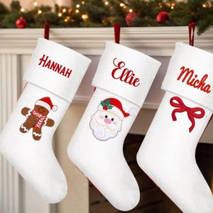 Peut inclure: Trois bas de Noël blancs avec une bordure rouge. Chaque bas porte un nom en écriture rouge : Hannah, Ellie et Micha. Les décorations incluent un bonhomme en pain d'épices, le Père Noël et un nœud rouge.