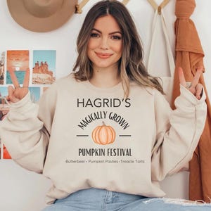 May include: A woman wearing a cream-colored sweatshirt with the text "Hagrid's Magically Grown Pumpkin Festival" and the words "Butterbeer - Pumpkin Pasties - Treacle Tarts" printed below. The sweatshirt features a cartoon pumpkin with a stem and leaves.