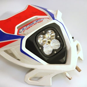 May include: White motorcycle headlight with a black housing and four LED lights. The headlight is mounted on a white plastic bracket with red and blue accents. The text "EXC Racing" is visible on the bracket.